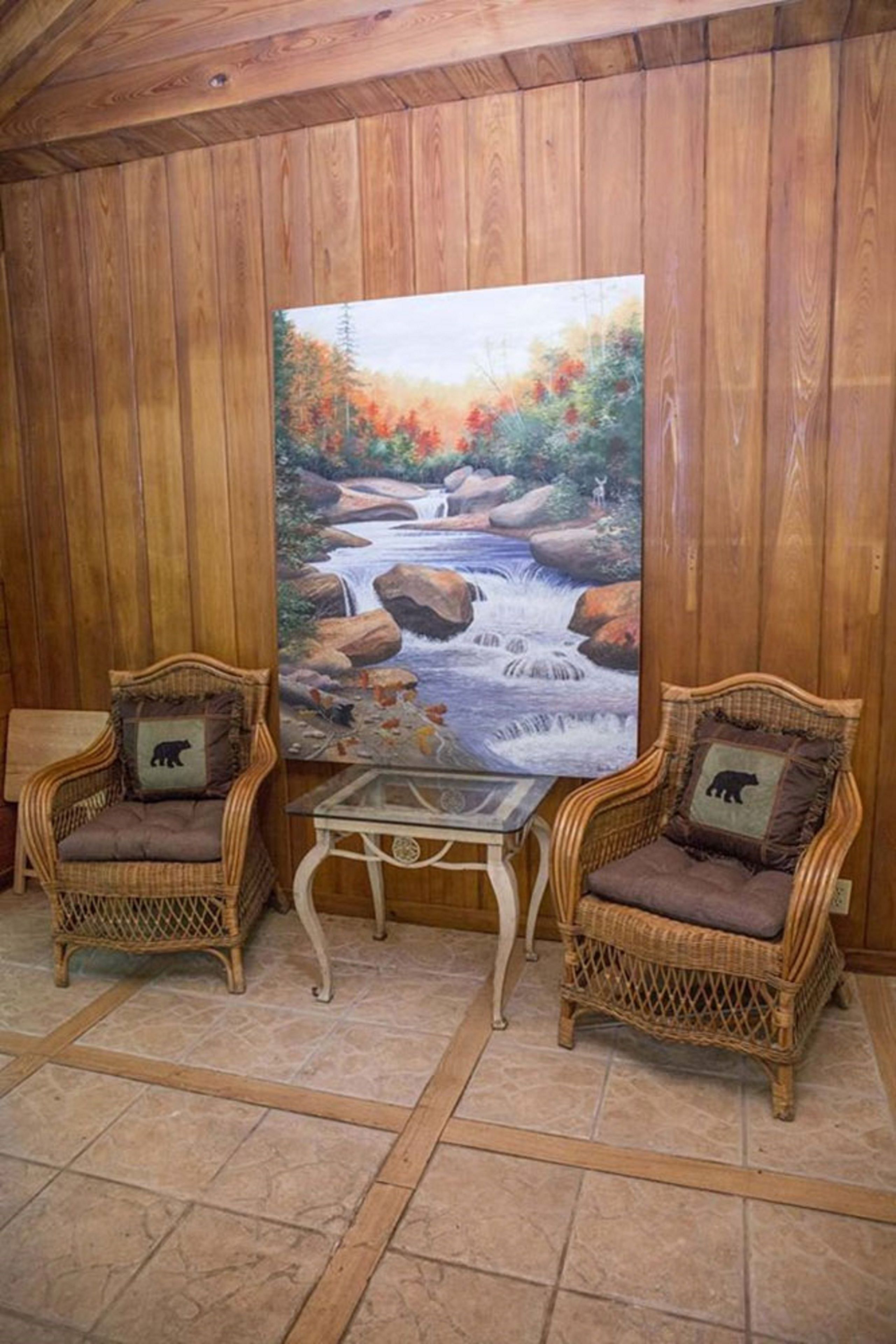 A room with wooden paneled walls, two wicker chairs with cushions, a glass table, and a painting of a river scene.