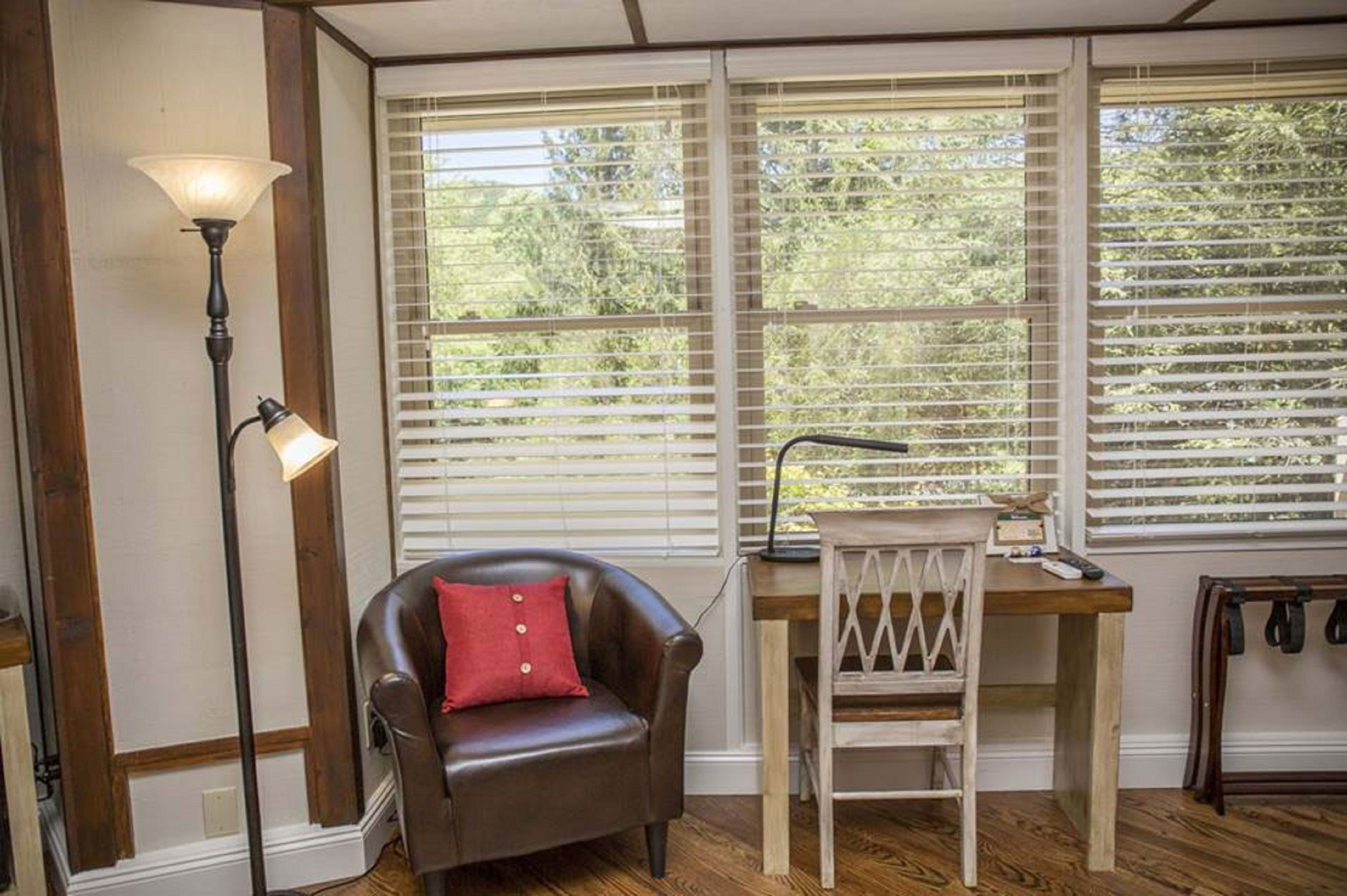 A room with a wooden floor, a leather armchair with a red pillow, a desk with a chair, and a floor lamp.