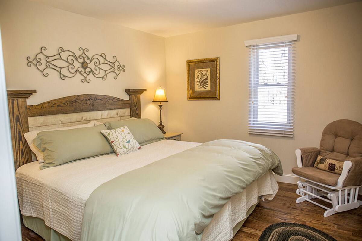 A bedroom with a large bed, a nightstand with a lamp, a rocking chair, and a window with blinds.