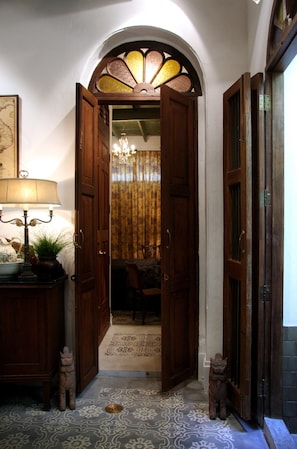 An open wooden door leading to a room with a patterned floor, a chandelier, and a lamp on a side table.