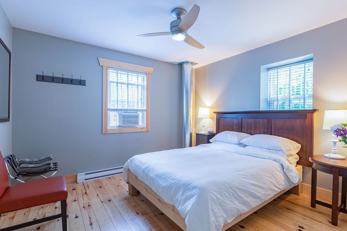 A bedroom with a bed, a ceiling fan, a window, a nightstand, a lamp, and a chair.