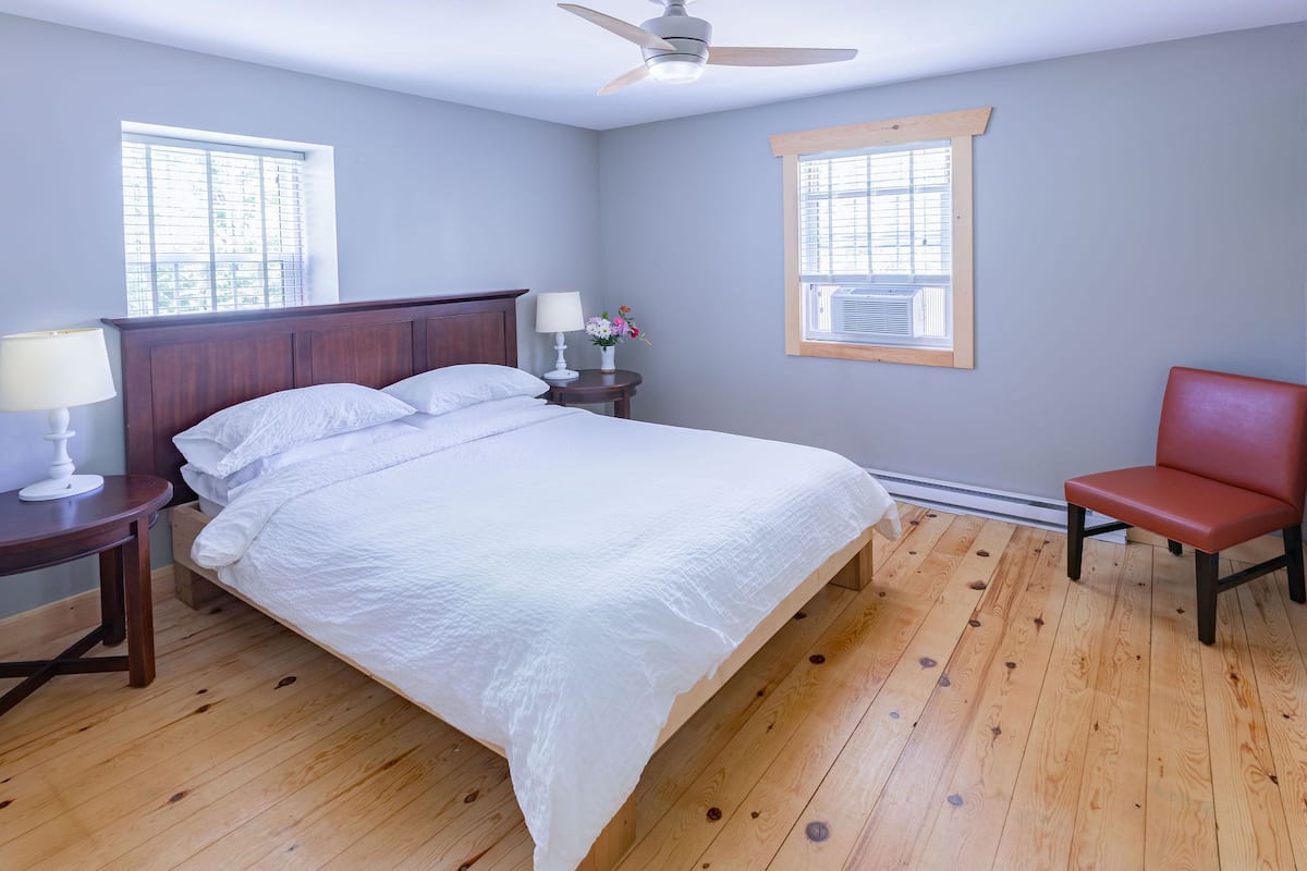 A bedroom with a bed, two bedside tables, a chair, and two windows.