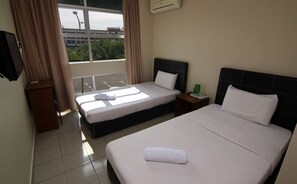 A hotel room with two beds, a window with curtains, a small table with a green cup, and an air conditioning unit on the wall.