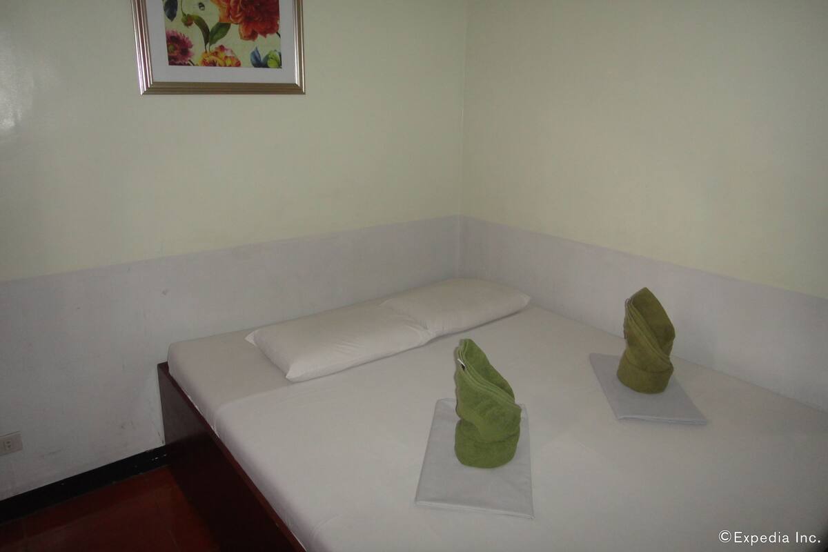 A single bed with white bedding and pillows, a green towel on a clear plastic bag, and a framed floral painting on the wall.