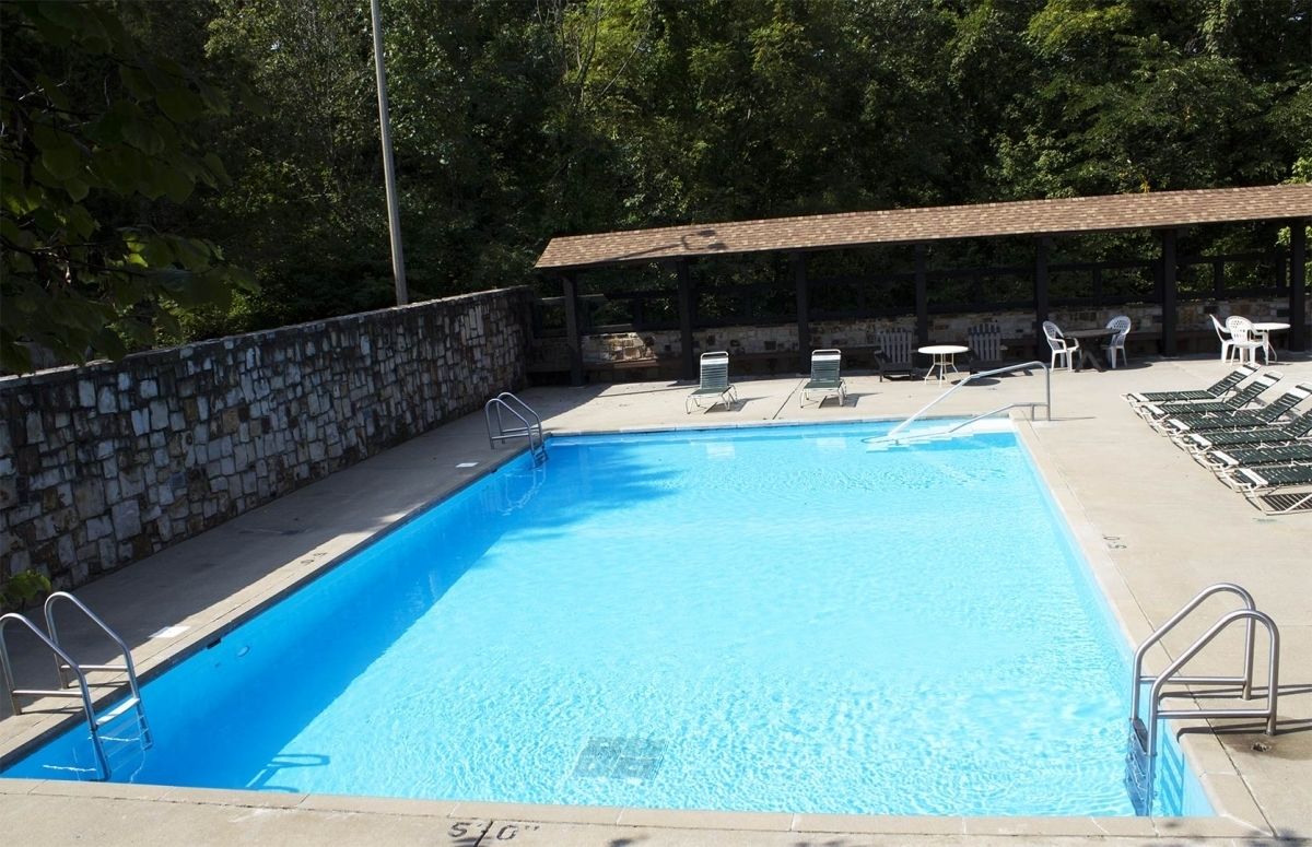 Una piscina rectangular con agua azul cristalina, rodeada por un muro de piedra y una zona cubierta con sillas y mesas.