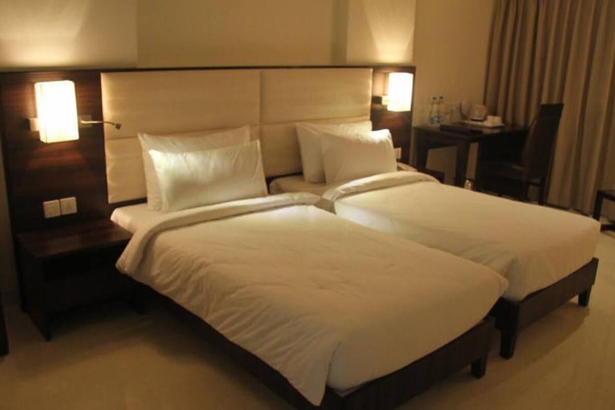 Una cama doble con dos almohadas, una mesita de noche con lámpara, una pequeña mesa con una silla y una ventana con cortinas.