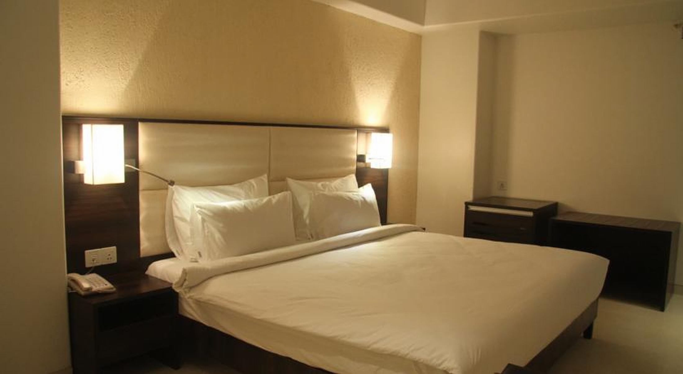 Quarto de hotel com cama, mesas de cabeceira, criado-mudo com telefone e luminária na parede.