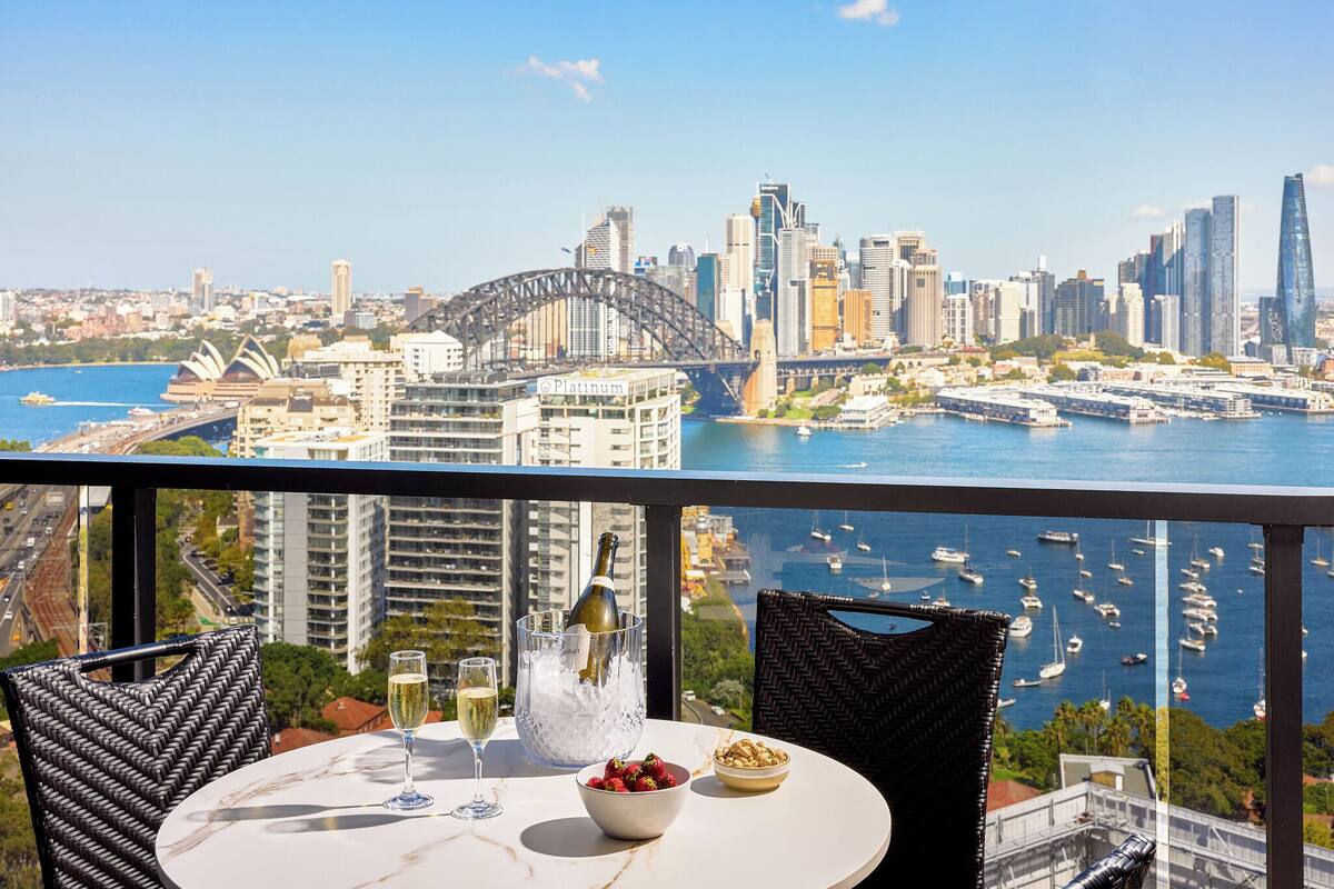 A balcony with a table, a bottle of champagne, a glass, a bowl of strawberries, and a view of the Sydney Opera House and Sydney Harbour.