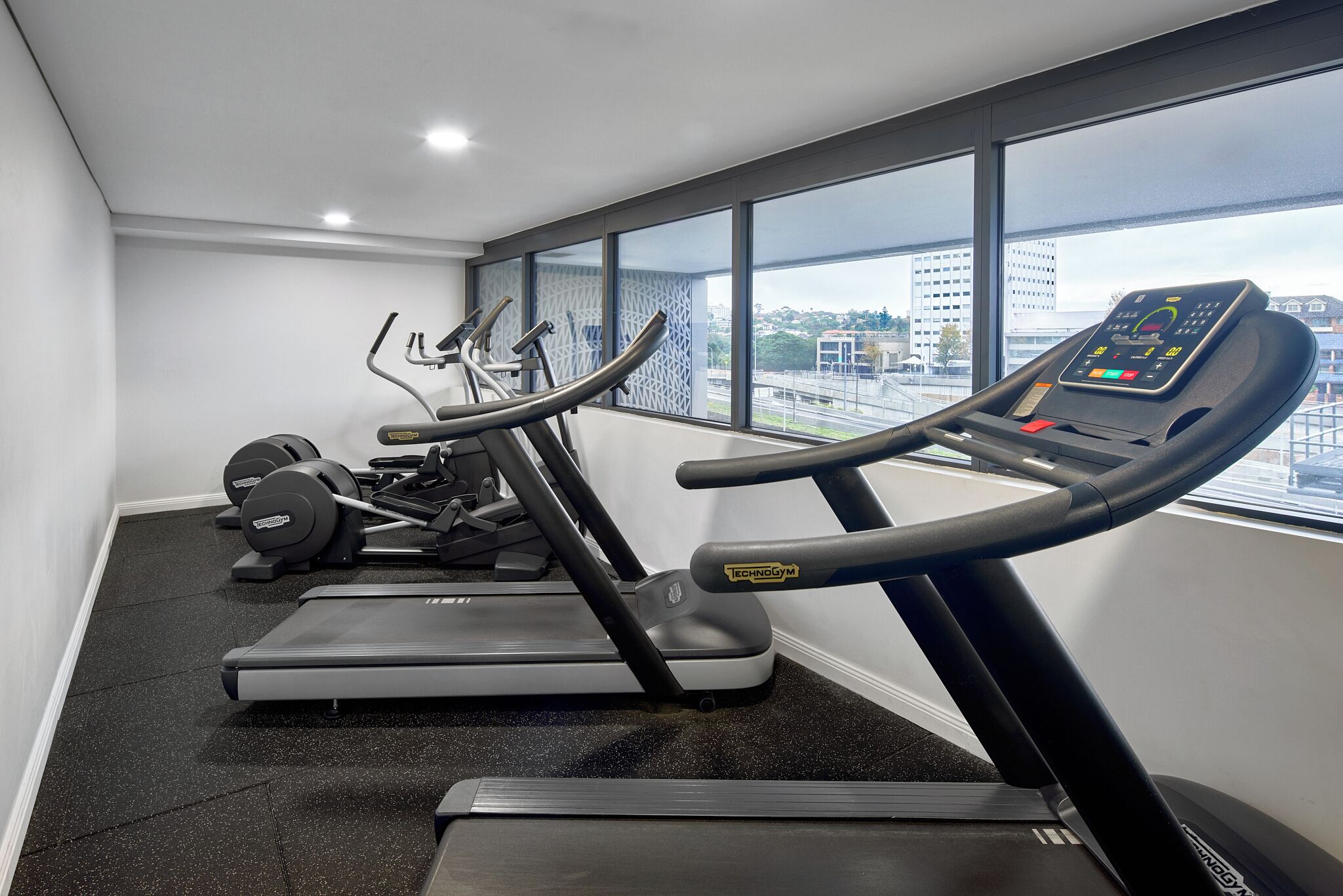 A gym with treadmills, ellipticals, and weight machines, large windows with a city view.