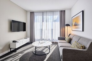 Una habitación de hotel moderna con una televisión de pantalla plana, una consola blanca, una mesa de centro redonda, un sofá gris y un ventanal con cortinas ligeras.