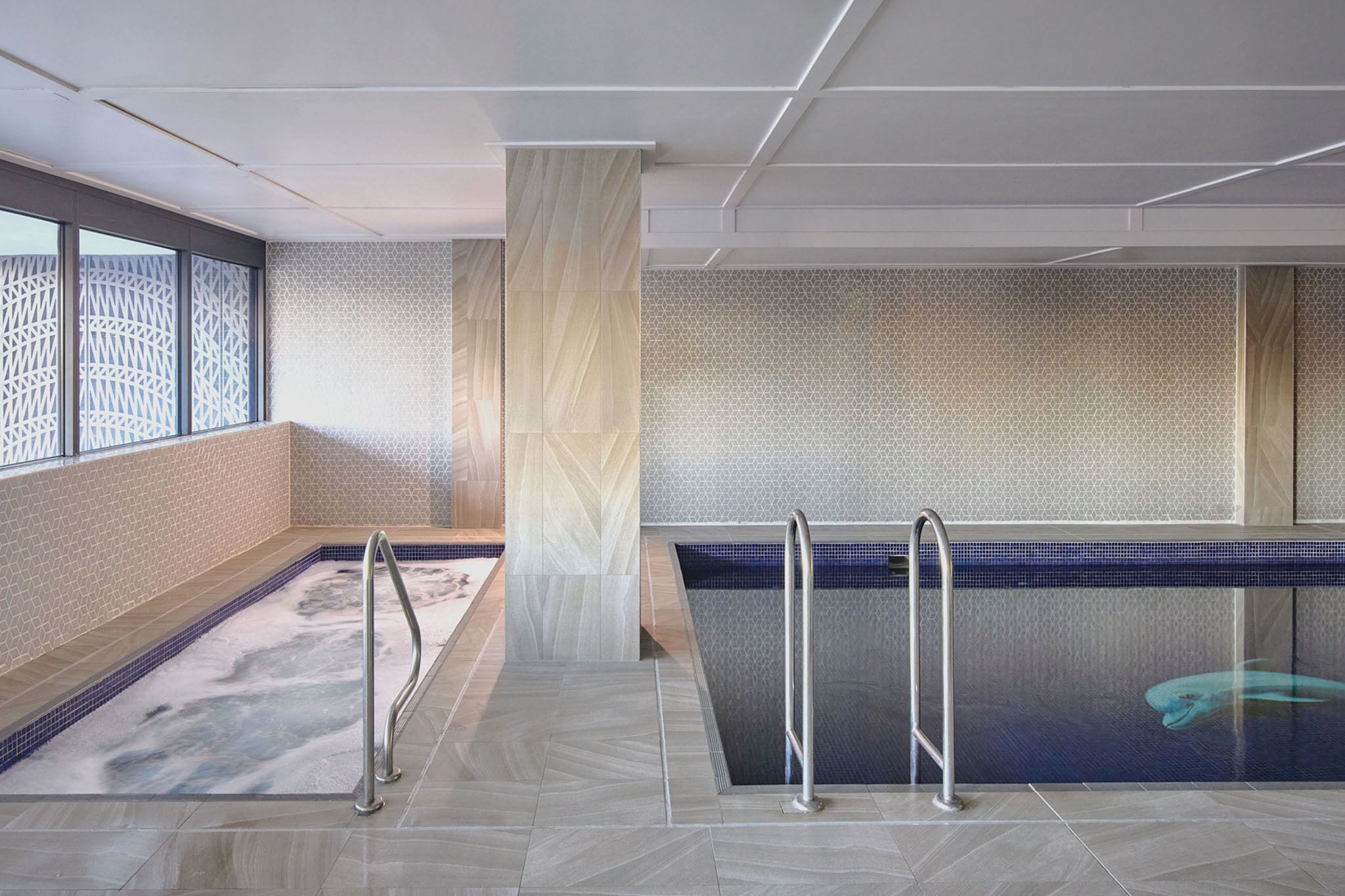 A modern indoor pool area with a geometric tile pattern, a central column, and metal handrails.