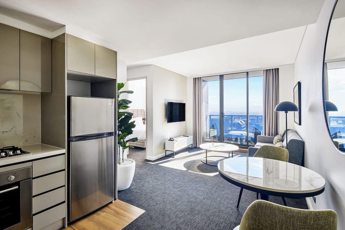 A modern hotel room with a kitchen, a dining area, a seating area with a view, and a large window.