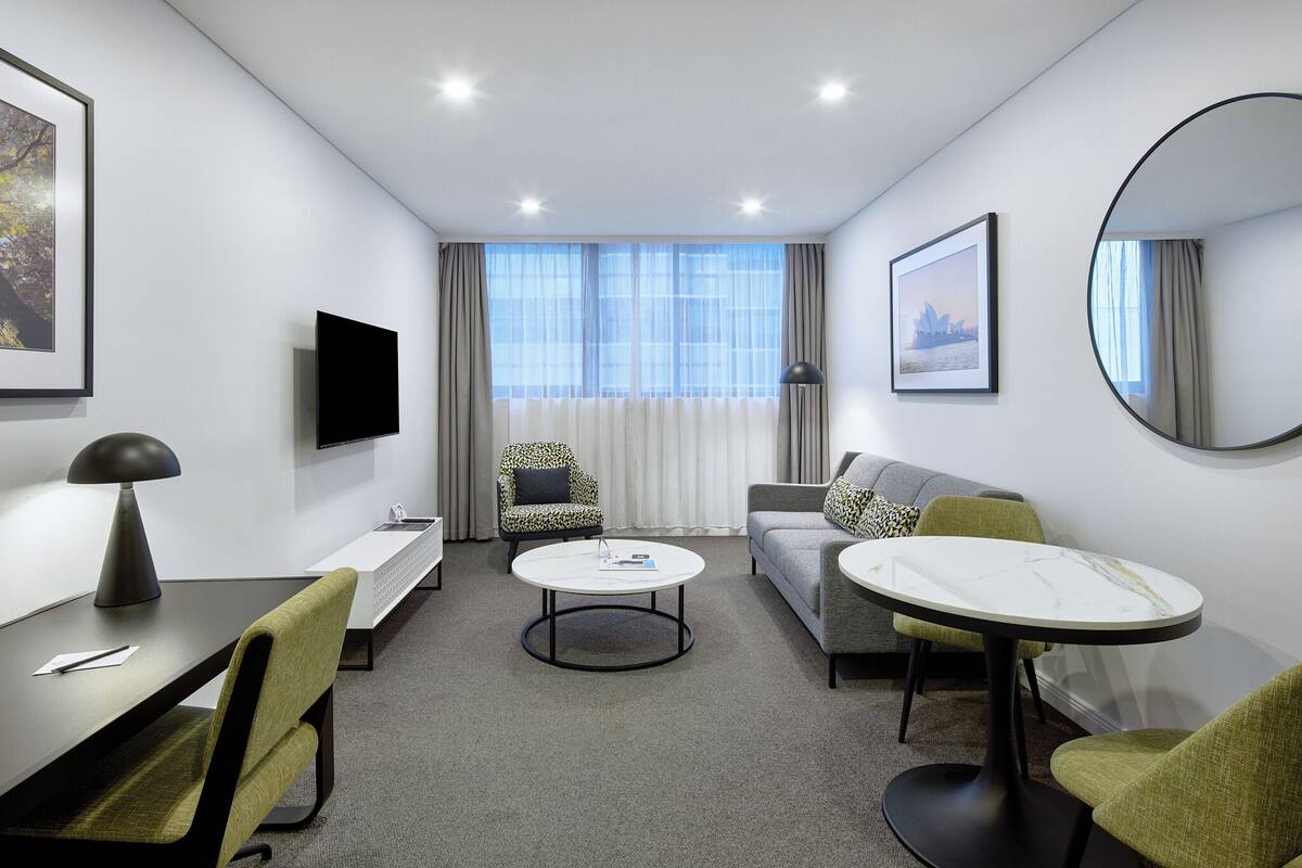 A modern hotel room with a sofa, a round dining table, a desk with a lamp, a TV, and a large window with curtains.