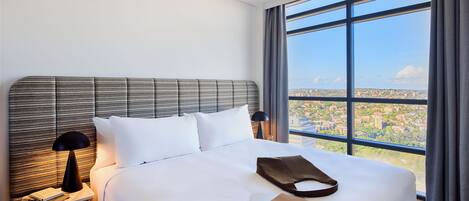 A hotel room with a large bed, a nightstand with a lamp, and a view of a cityscape through large windows.