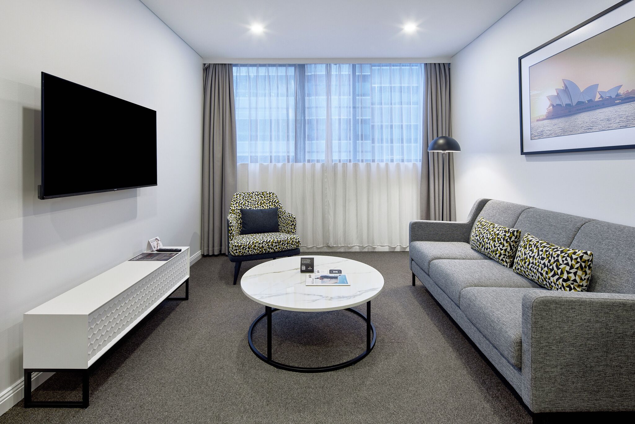 A modern hotel room with a flat-screen TV, a sofa, a patterned armchair, a coffee table, and a framed picture of a famous landmark.