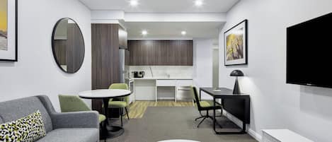 Una habitación de hotel moderna con un sofá gris, una mesa de centro redonda, una zona de comedor con una mesa y sillas blancas, un área de cocina y un televisor de pantalla plana montado en la pared.
