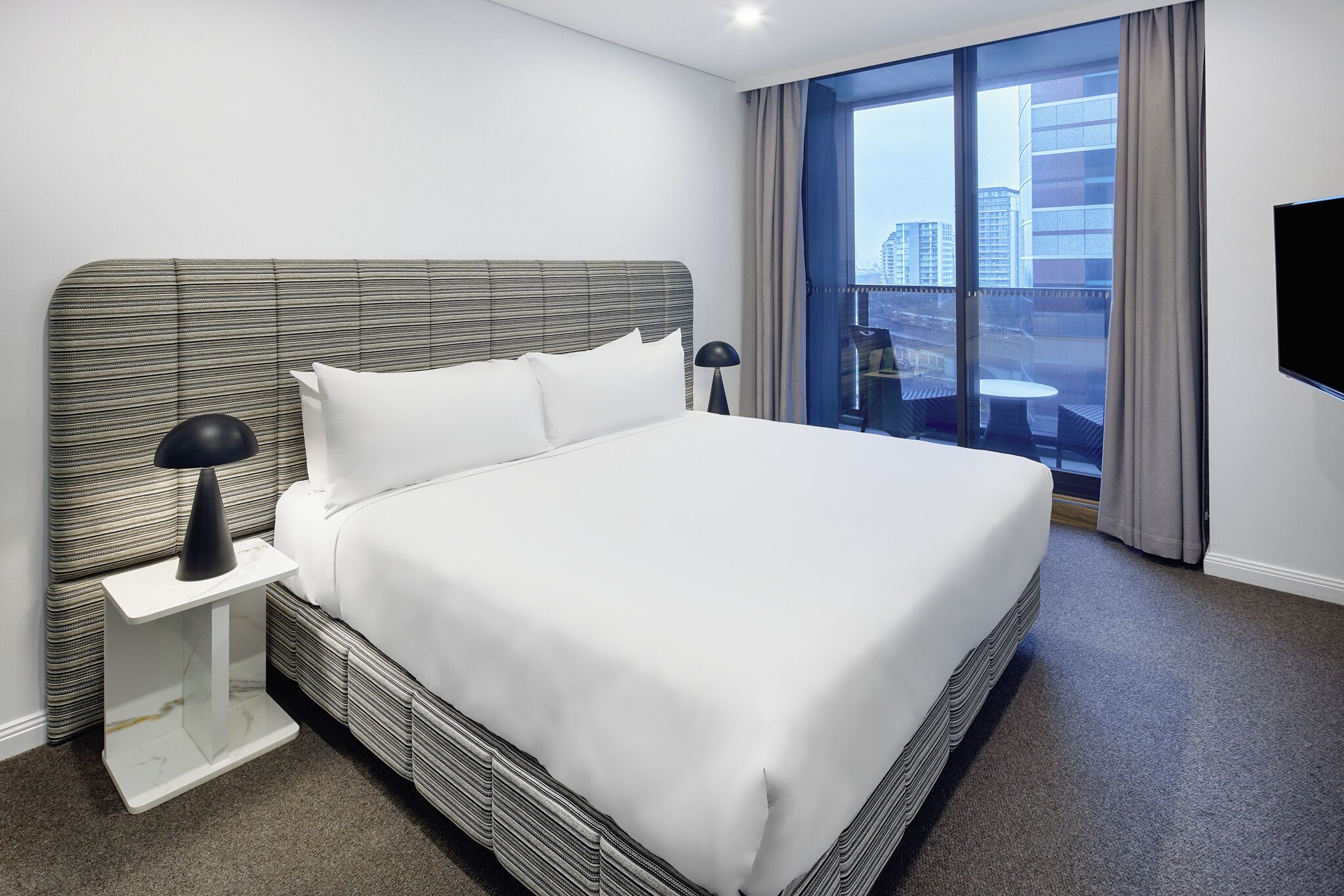 A hotel room with a large bed, a nightstand, a lamp, a TV, and a balcony with a city view.