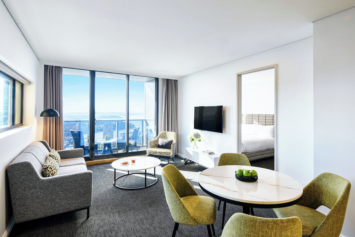 A modern hotel room with a large window, a sofa, a dining table, and a TV.