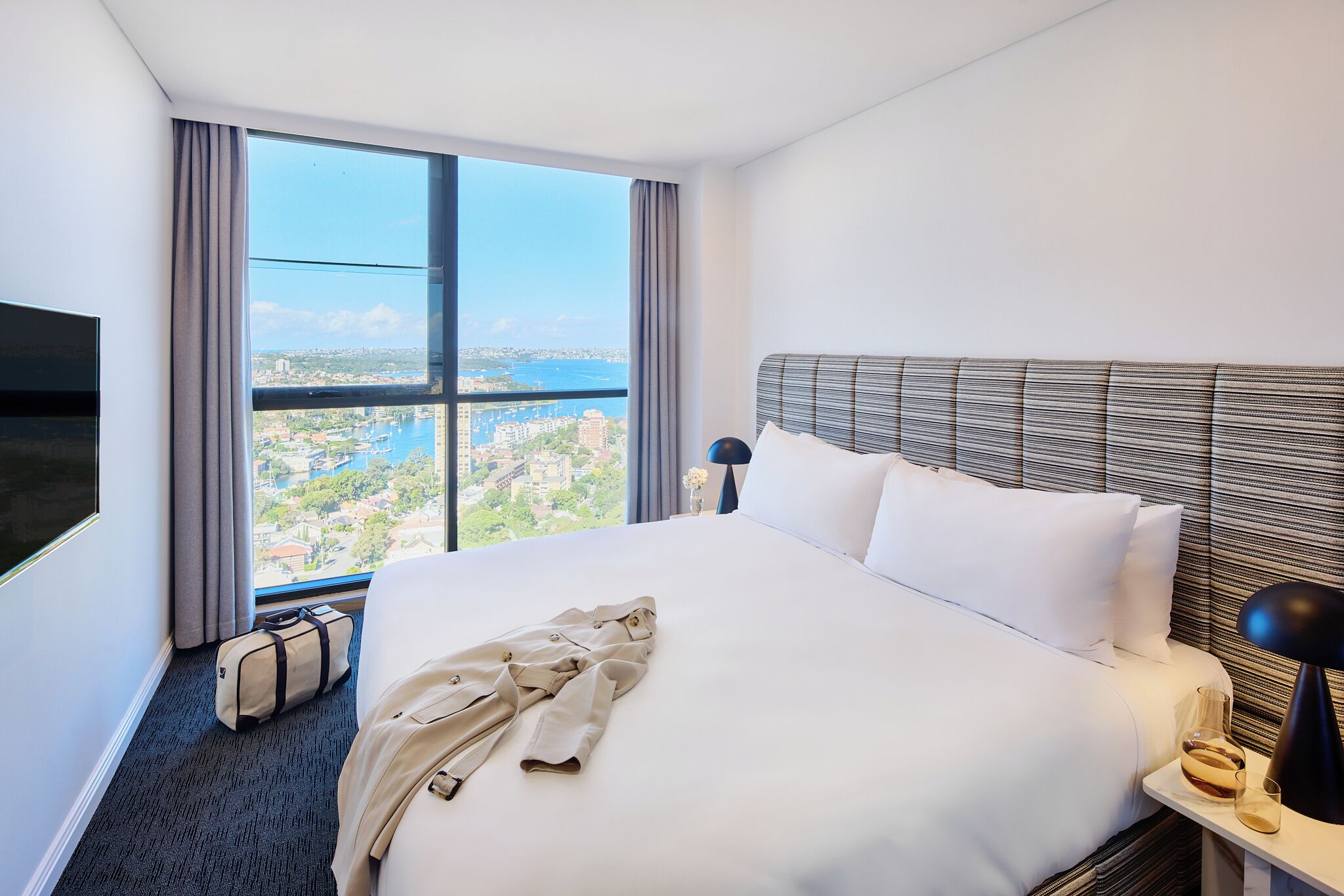 Une chambre d’hôtel avec un grand lit, une télévision, une table de chevet, une lampe et une vue sur le paysage urbain, visible à travers une grande fenêtre.