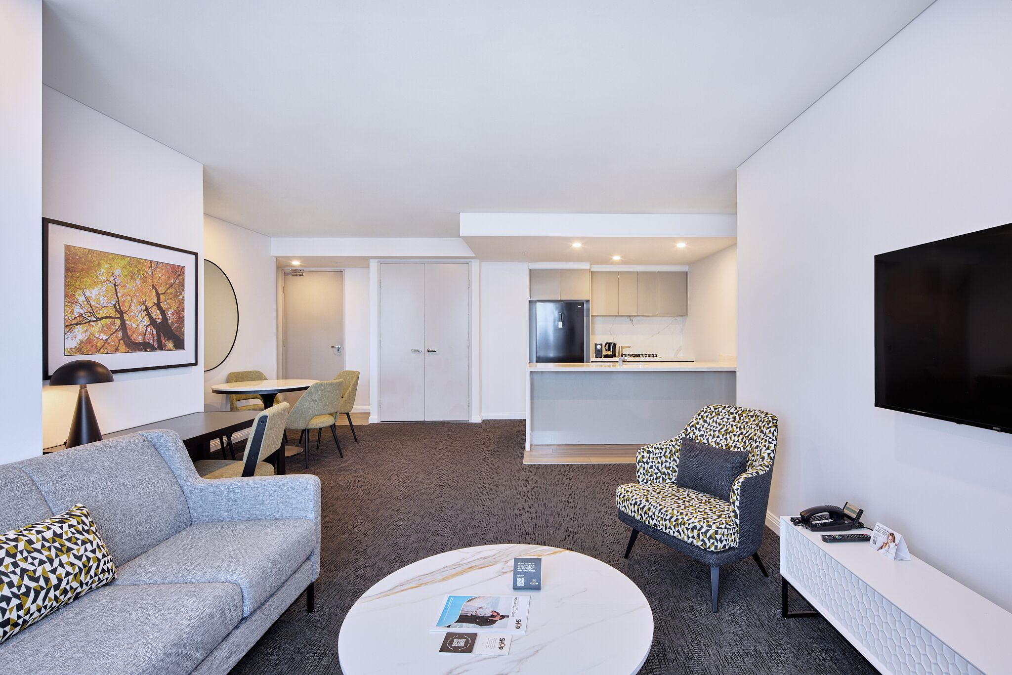 A modern hotel room with a sofa, armchair, dining area, kitchen, and a flat-screen TV.