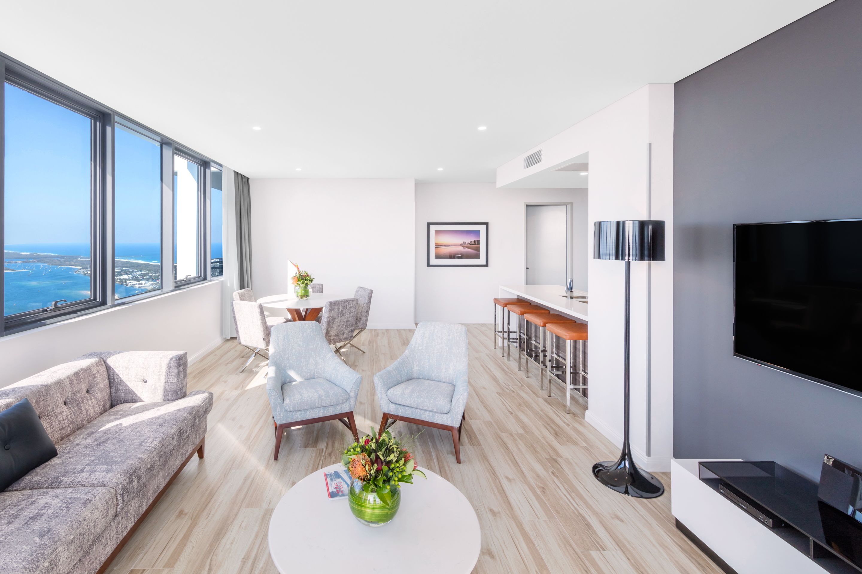 A modern living room with a large window, a sofa, two armchairs, a coffee table with a vase, a flat-screen TV, and a bar area.