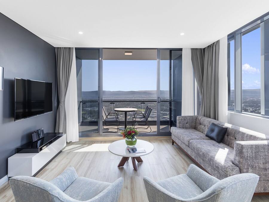 A modern living room with a flat-screen TV, grey sofas, and a balcony with a city view.