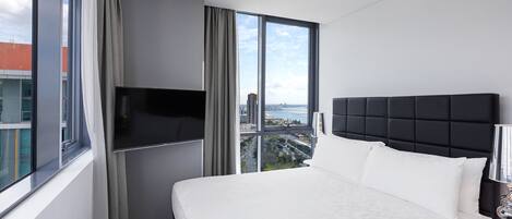 A modern hotel room with a large bed, a flat-screen TV, and a city view through large windows.