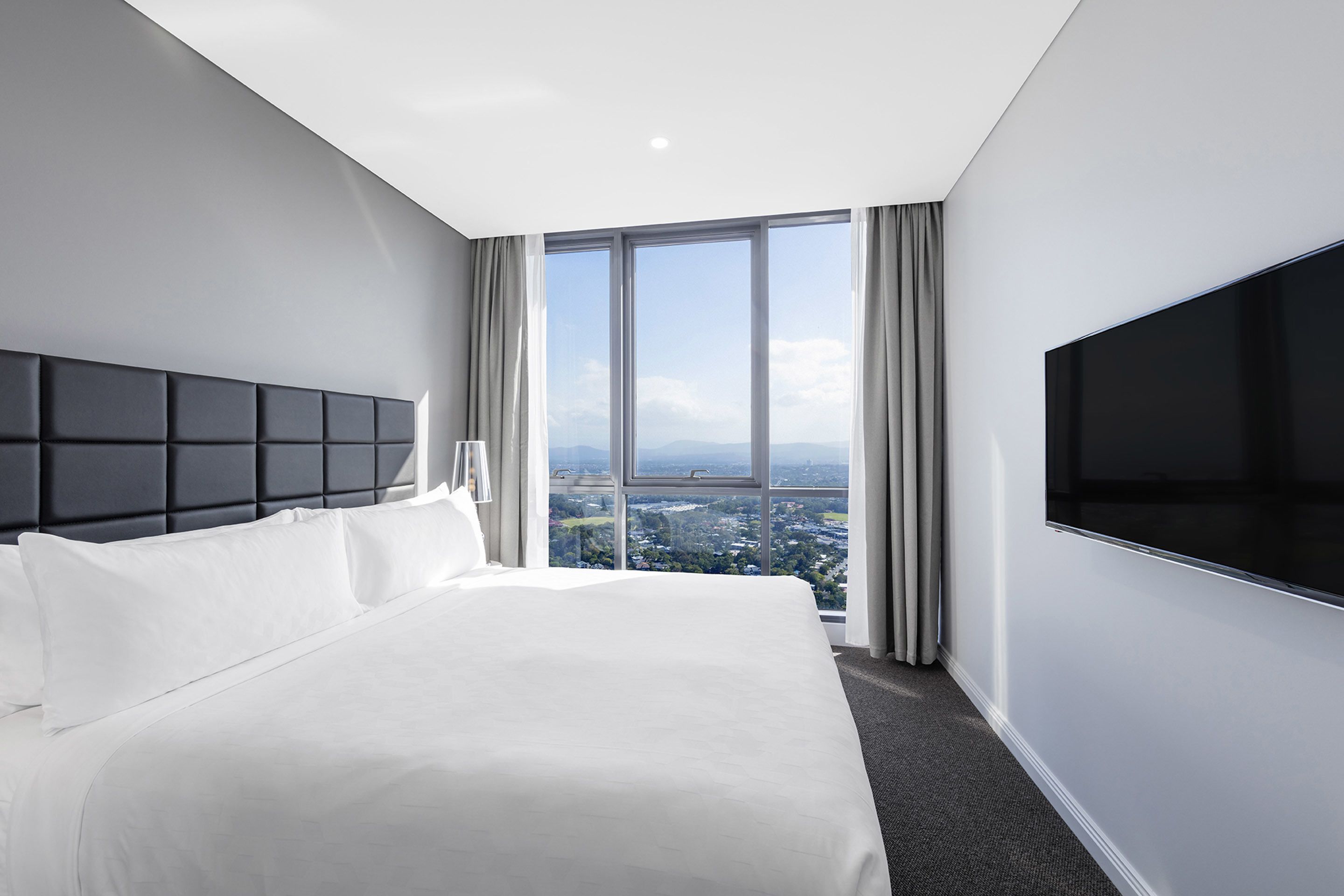 A modern hotel room with a large bed, a flat-screen TV, and a city view through large windows.