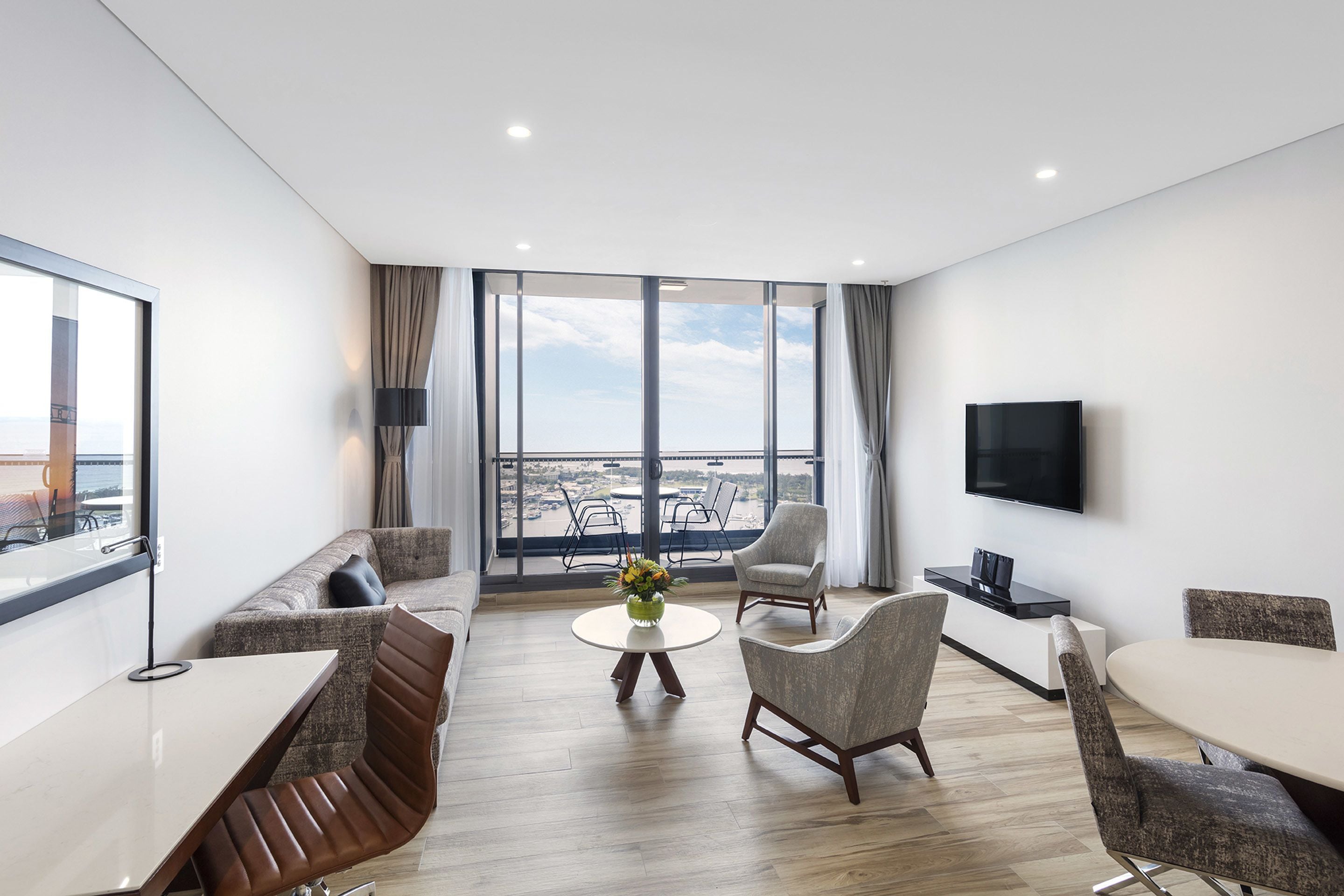 A modern hotel room with a balcony, a flat-screen TV, a dining area with a round table and chairs, and a sofa.