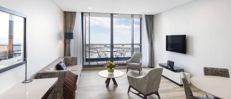 A modern hotel room with a balcony, a flat-screen TV, a dining area with a round table and chairs, and a sofa.