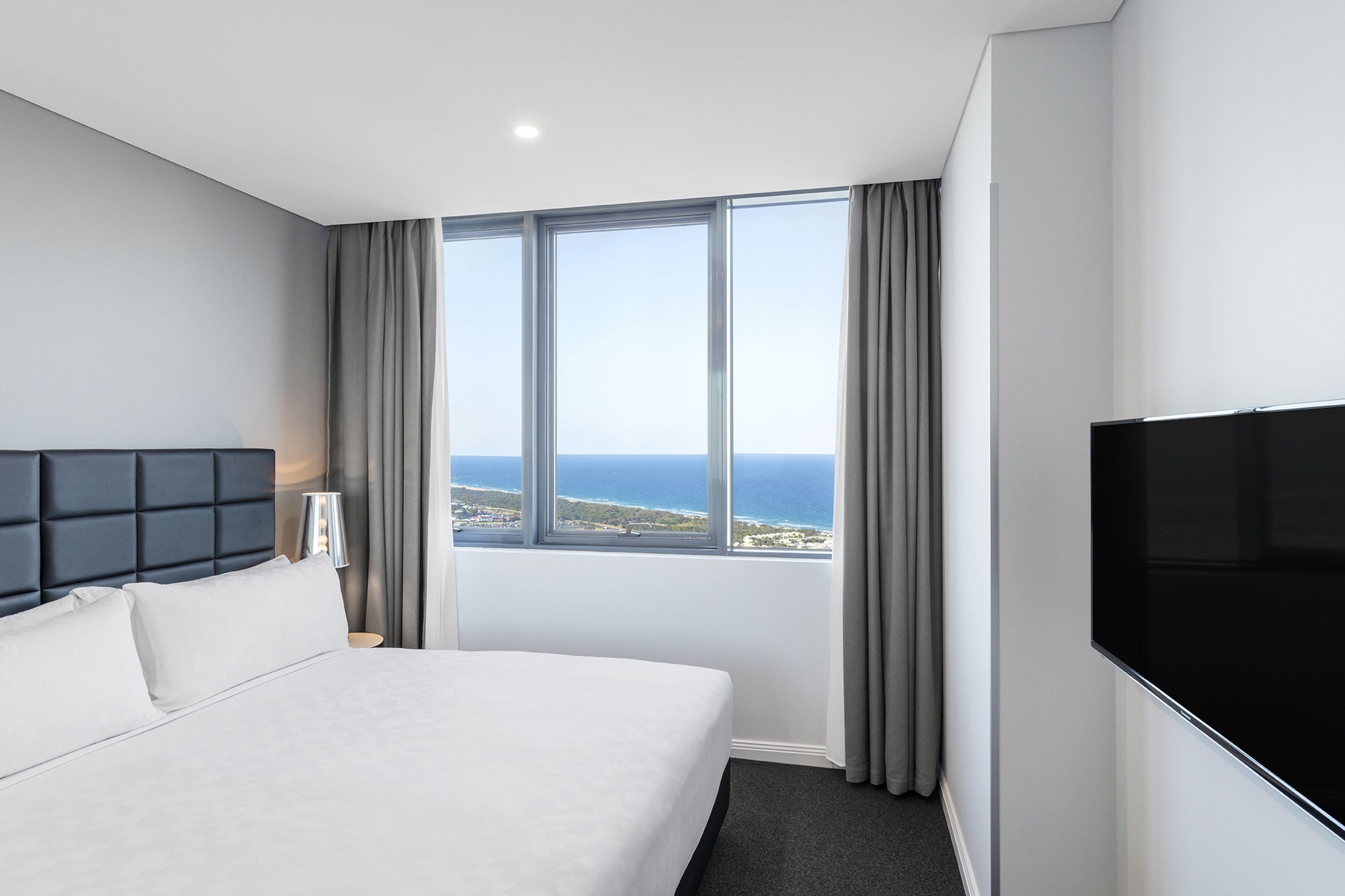 Quarto de hotel com uma cama grande, televisão de tela plana, janela com vista para o oceano e cortinas cinzas.