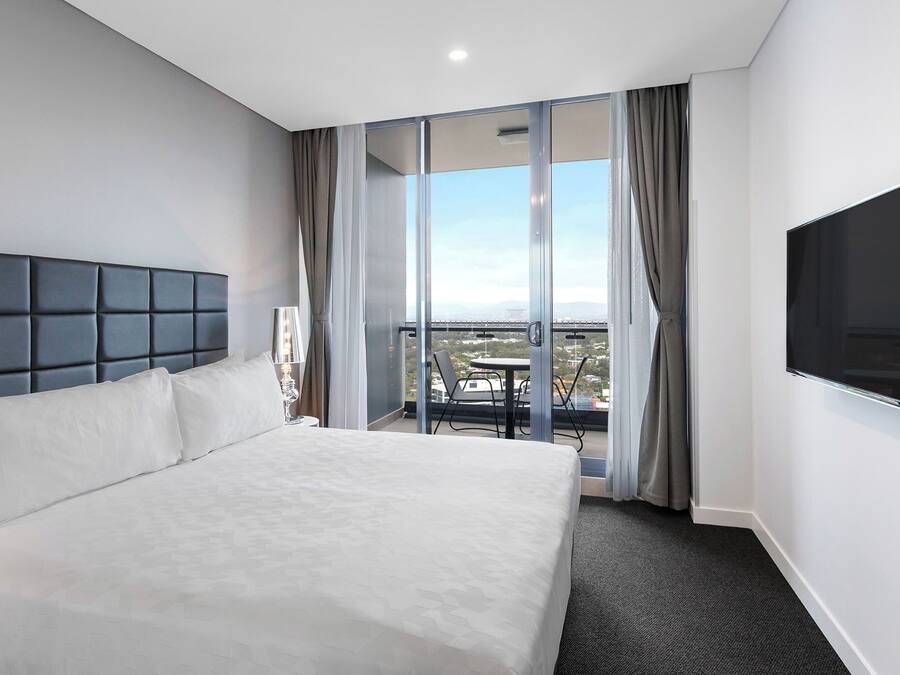 A hotel room with a large bed, a flat-screen TV, a balcony with a table and chairs, and a city view.
