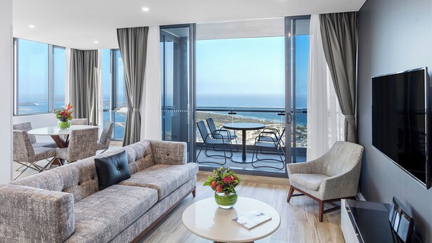 Een moderne woonkamer met een bank, fauteuils, een salontafel en een televisie. Er zijn grote ramen met uitzicht op de oceaan.