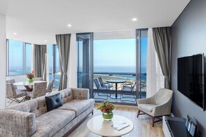 Un salon moderne comprenant un canapé, des fauteuils, une table basse et un téléviseur. De grandes fenêtres offrent une vue sur l’océan.