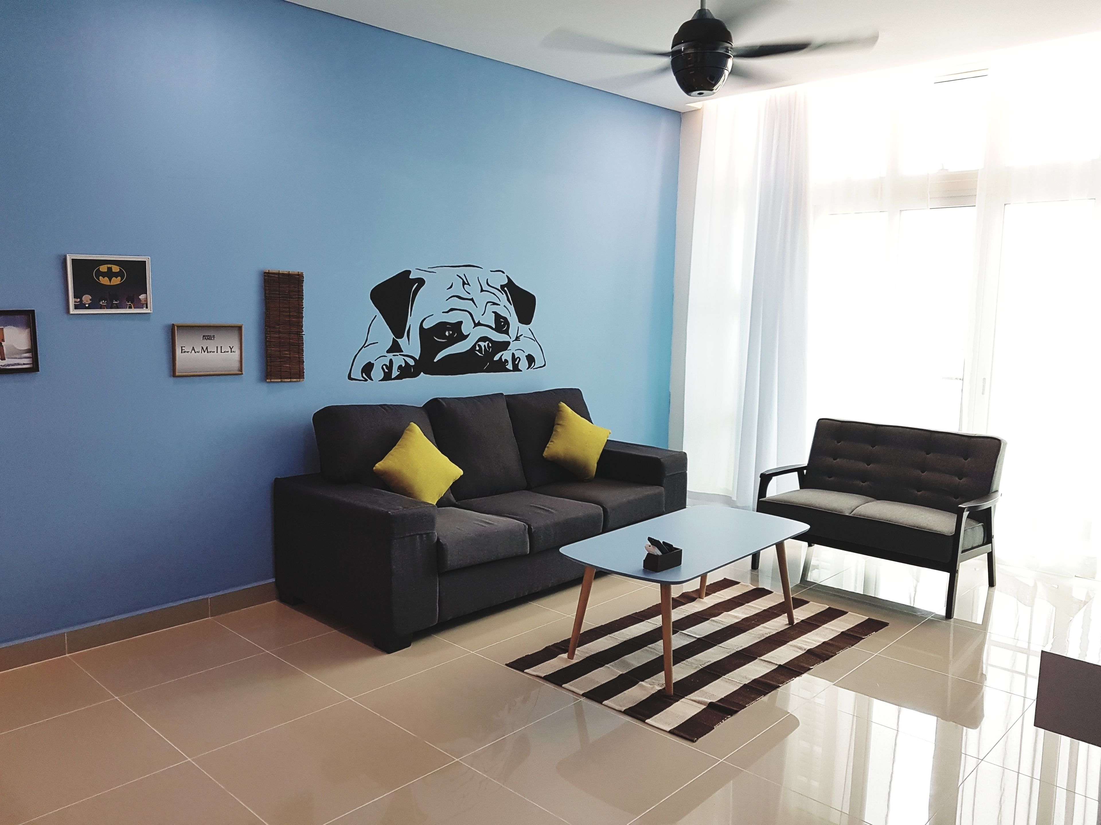 A modern living room with a blue accent wall, a black sofa with yellow cushions, a black armchair, a glass coffee table, and a ceiling fan.