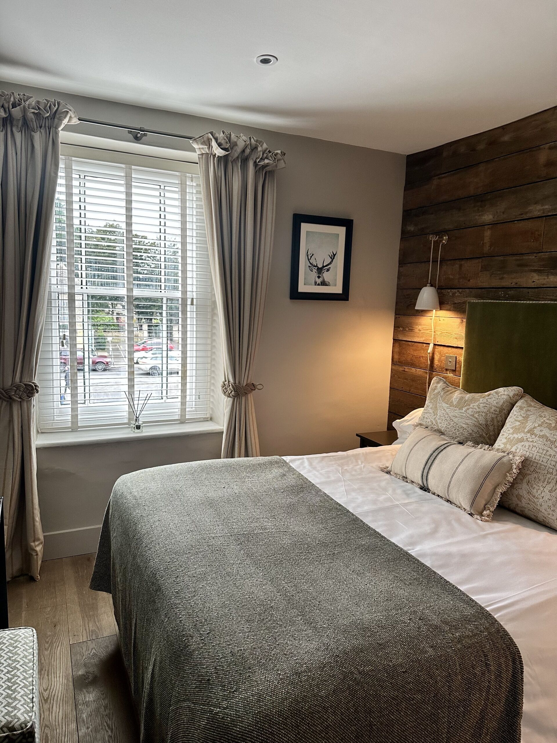 A bedroom with a large bed, a window with curtains, a framed picture on the wall, and a bedside lamp.