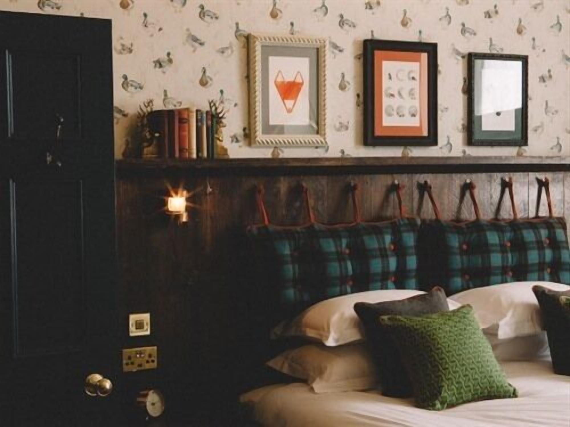 A bedroom with a bed, pillows, a wall-mounted shelf with books and decorative items, and a door with a handle.