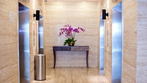 A modern elevator with marble walls, a console table with a vase of purple flowers, and a trash bin. - Vouk Hotel Suites, By The Blanket (George Town)