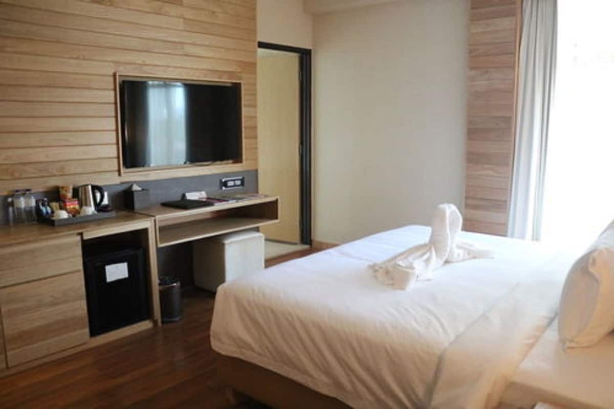 A hotel room with a bed, a desk with a television, a small trash bin, and a window with curtains.