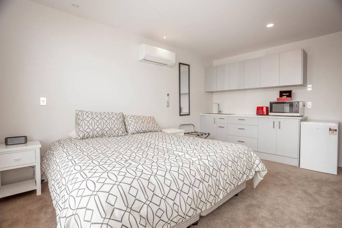 Een slaapkamer met een groot bed, een nachtkastje, een klein bureau, een kledingkast en een kitchenette met een magnetron en een koelkast.