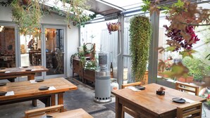 Un espacio al aire libre con mesas y bancos de madera, rodeado de plantas colgantes y macetas.