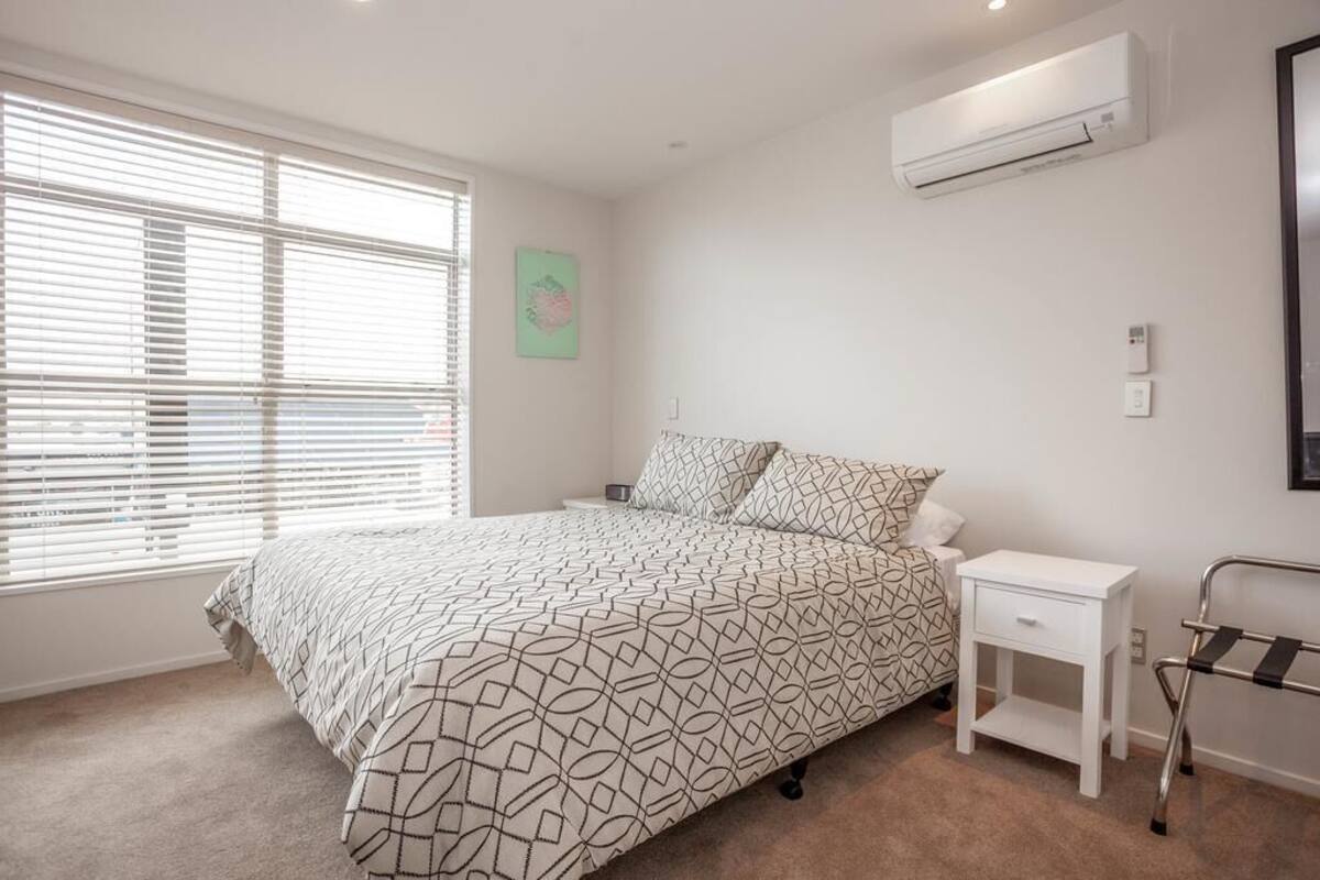 Een slaapkamer met een bed, nachtkastje, airconditioning, raam met jaloezieën en een wandlamp.