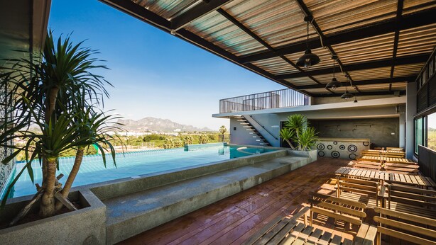 A poolside area with wooden benches, a concrete planter with a palm tree, and a view of a mountain range. - C Cha-am Hotel (Cha-am)