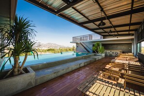 Un espace aménagé au bord de la piscine, avec des bancs en bois, un bac à fleurs en béton où est planté un palmier, et une vue sur une chaîne de montagnes.