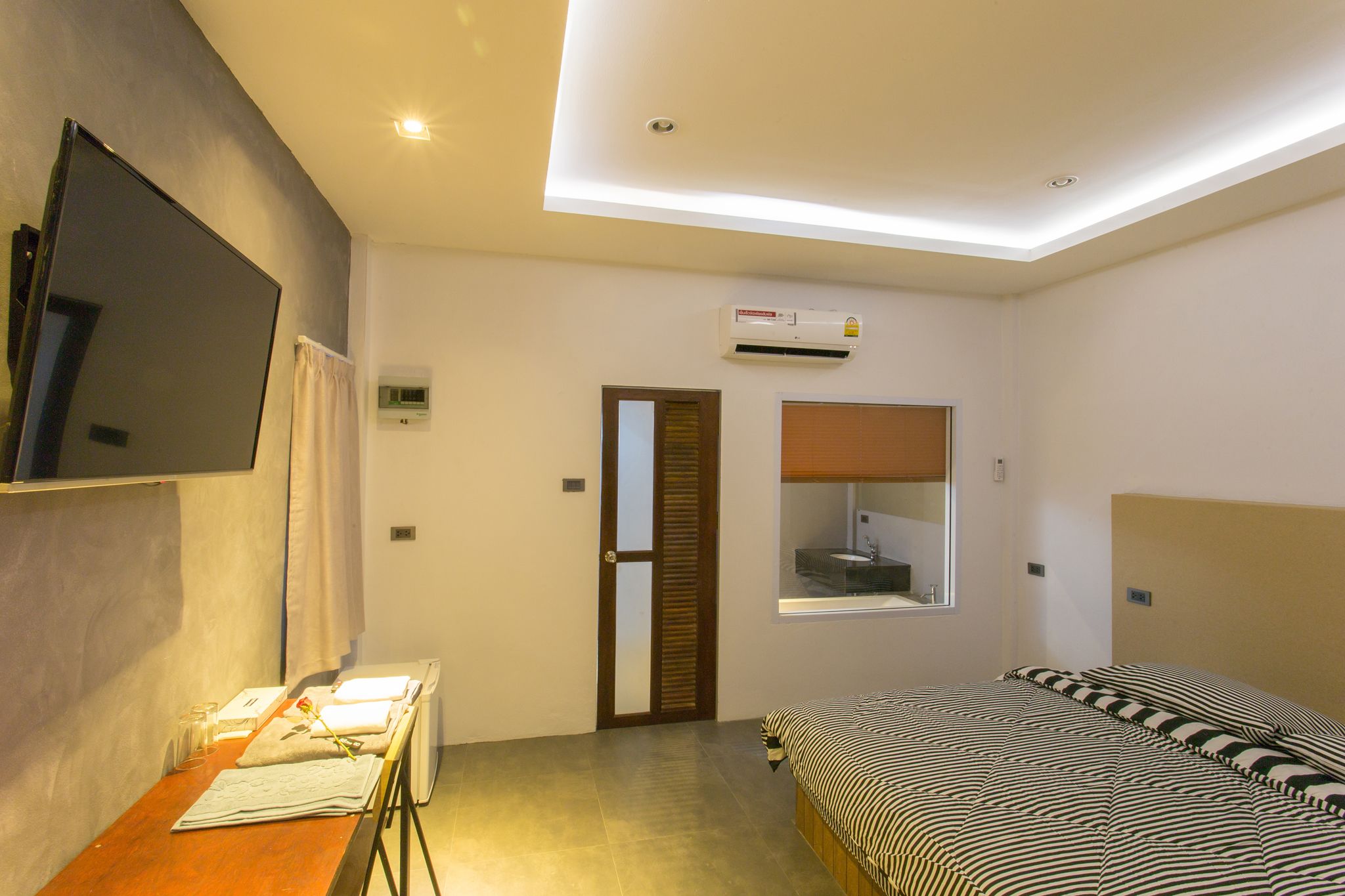A hotel room with a bed, a desk with a chair, a flat-screen TV mounted on the wall, an air conditioning unit, and a window with a view of the outside.