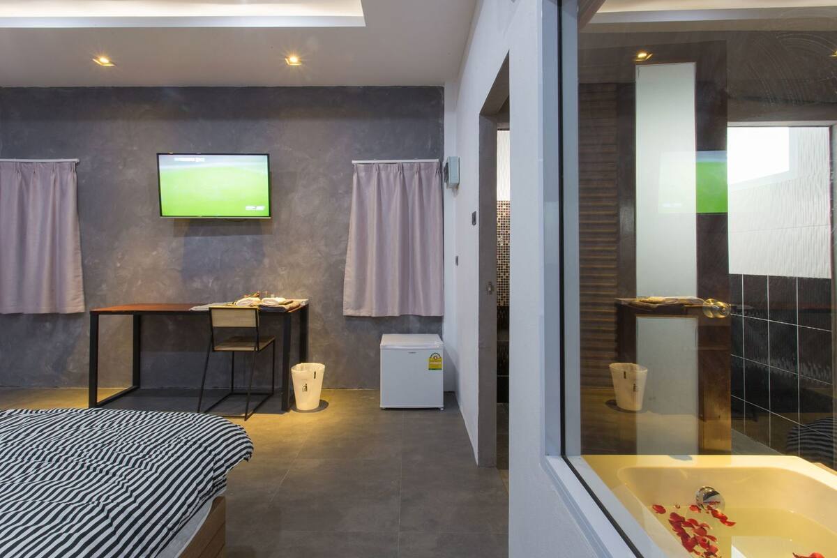 A modern hotel room with a bed, a desk, a television, a small refrigerator, and a bathroom with a bathtub.