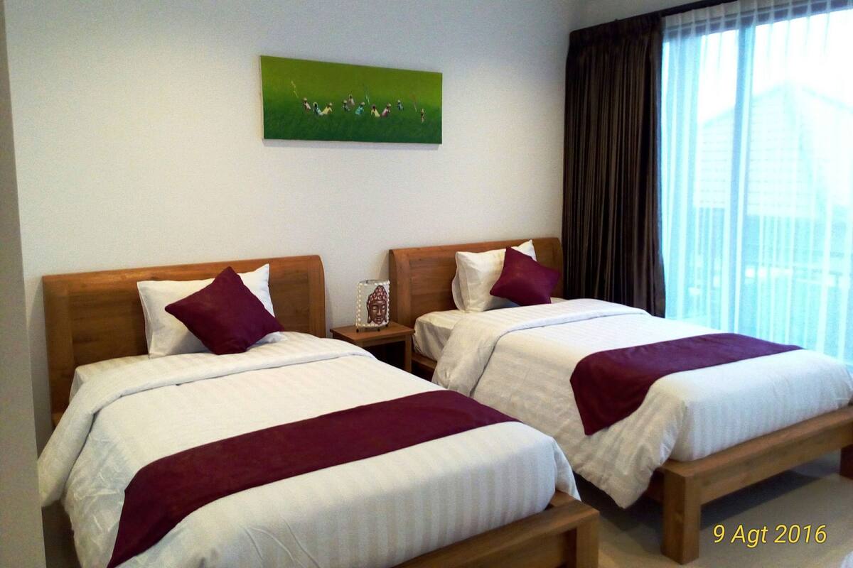 Quarto de hotel com duas camas, cada uma com cabeceira de madeira, roupa de cama branca e manta vinho. Um Buda está sobre a mesa de cabeceira, e um quadro com paisagem verde na parede.