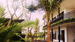 Un edificio moderno con un balcón, rodeado de abundante vegetación, incluyendo palmeras y otras plantas tropicales.