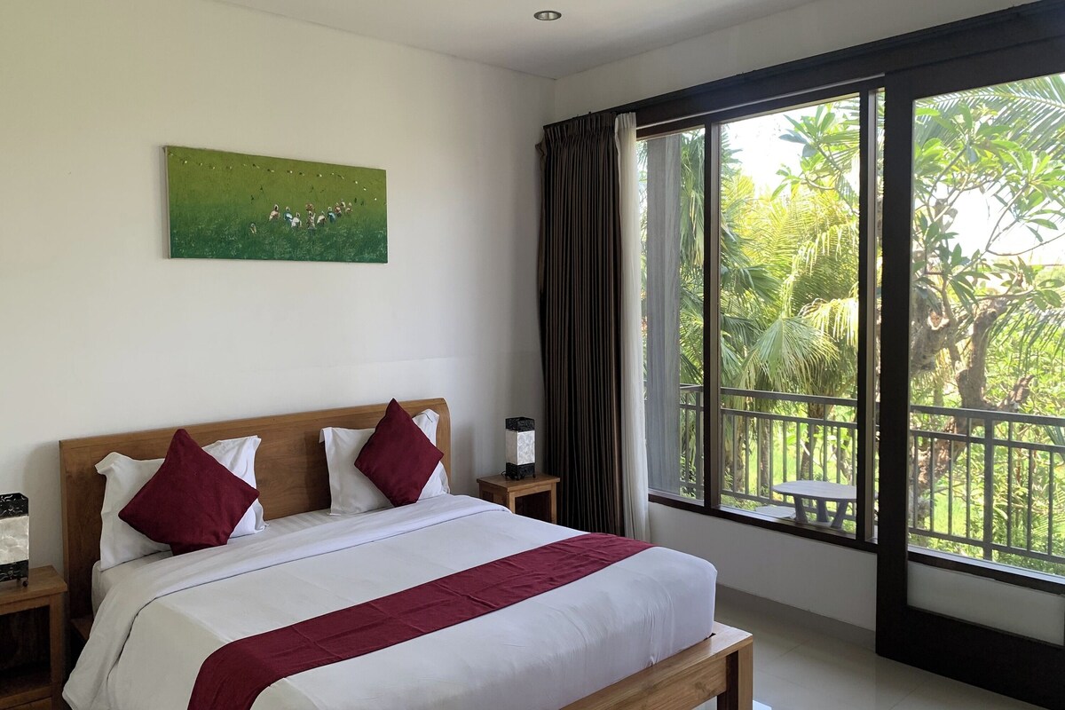 A hotel room with a large bed, a nightstand, a painting on the wall, and a window with a view of greenery.