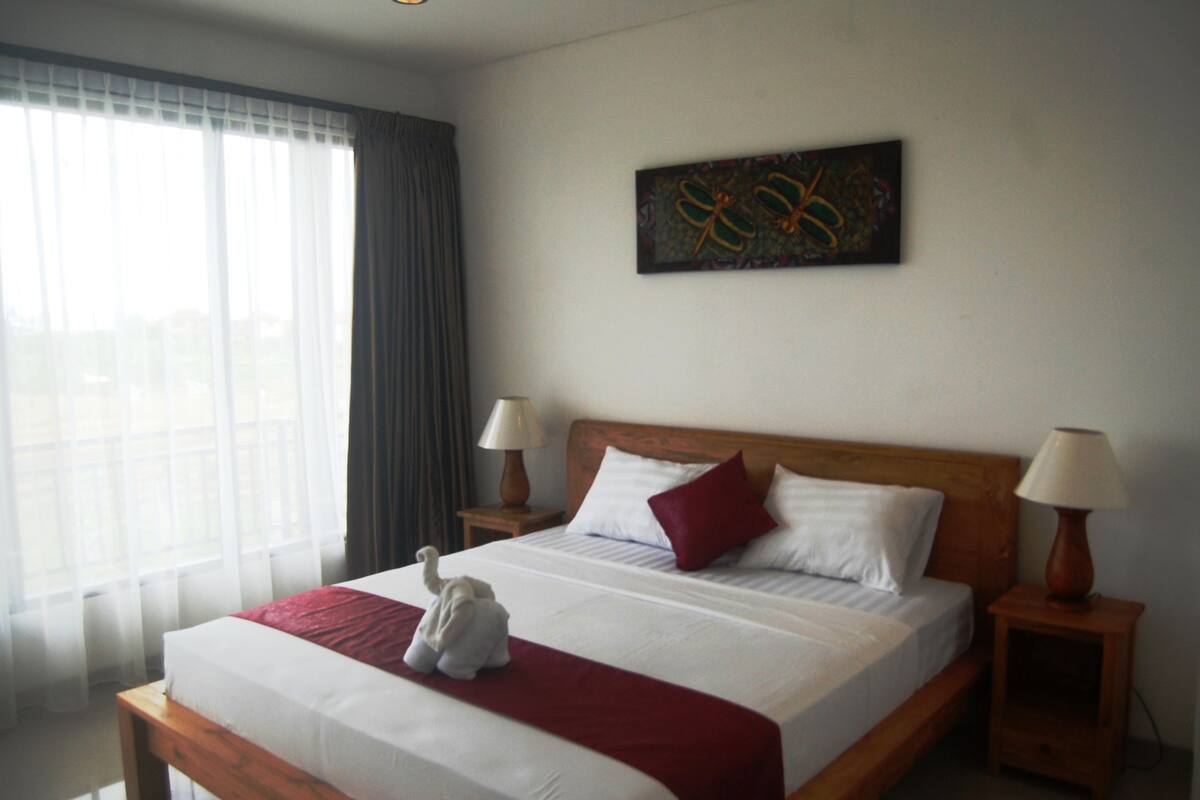 Quarto de hotel com uma cama grande, duas mesas de cabeceira, uma janela com cortinas e um quadro pendurado na parede.